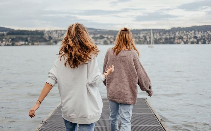 two women walk to the edge of a dock, in a body of water, looking toward the city on the other shore